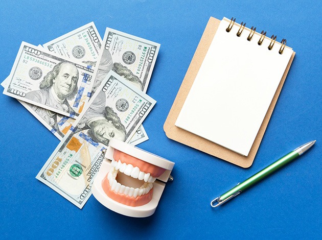 Bird’s eye view of model teeth next to $100 bills, notepad, and penil on blue surface